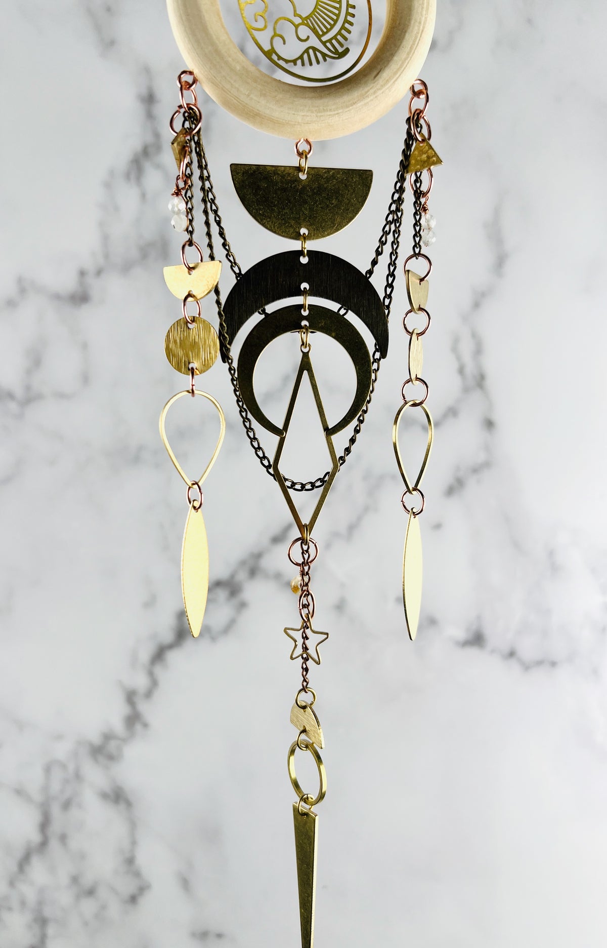 The bottom of a light wood ring with a center Charm of Sun and clouds, connected to the wood, are various metal items, including; bronze chain, gold colored, half circles, circle, circles, teardrop shapes, stars, circles and crescent moons as well as moonstone and citrine wire wrapped beads. The piece hangs in front of a marble wall.