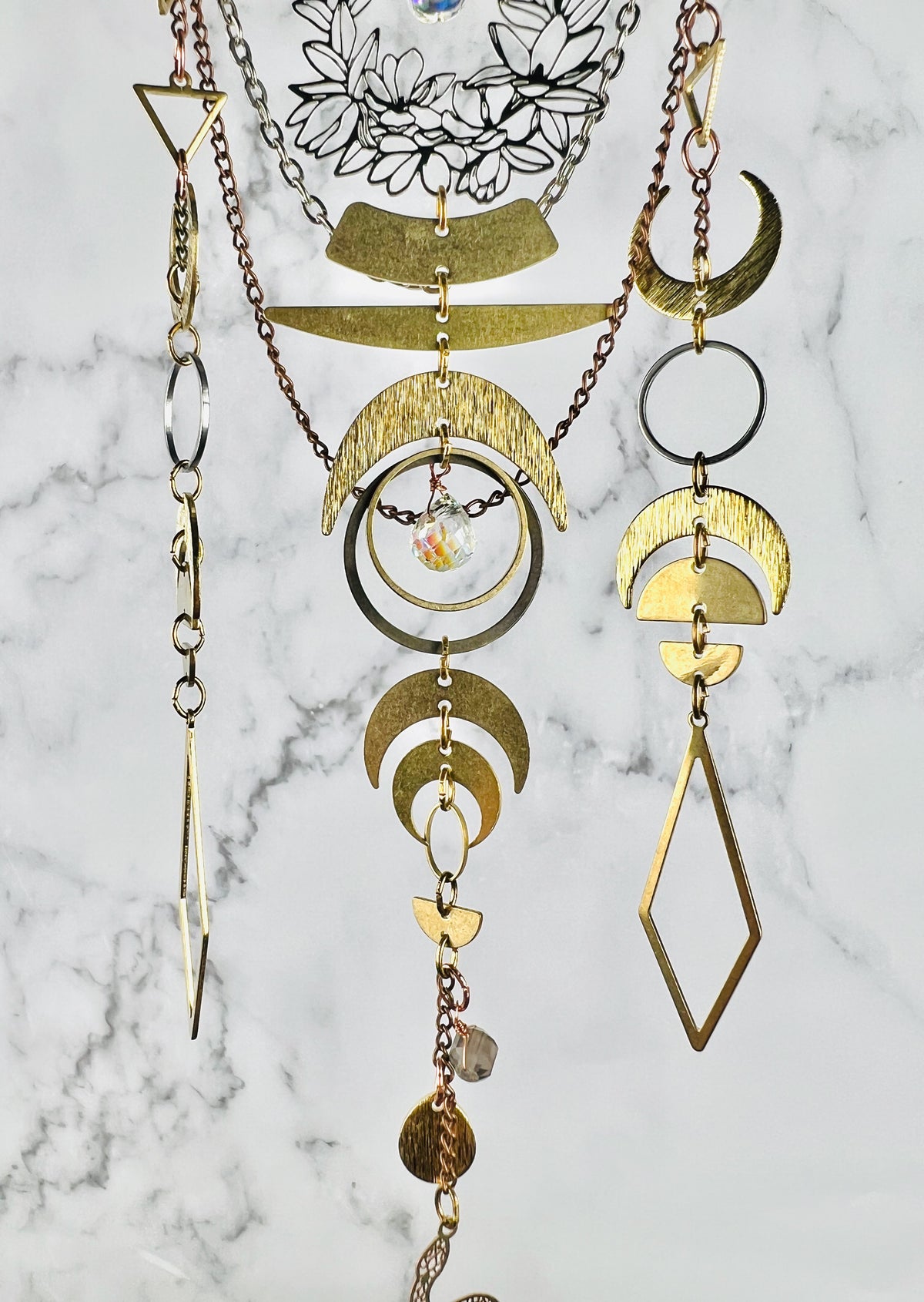 A layered bohemian-style metal crystal art for hanging on the wall, featuring gold colored metal crescent moon, geometric shapes, diamonds, a. prism and wire wrapped iolite beads and circular elements, displayed against a gray and white marble background.
