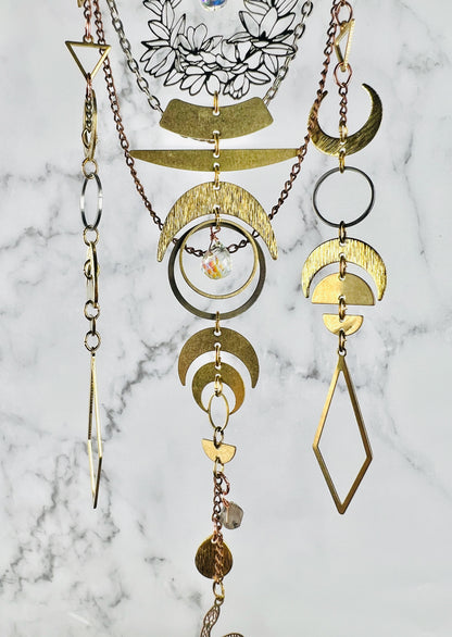 A layered bohemian-style metal crystal art for hanging on the wall, featuring gold colored metal crescent moon, geometric shapes, diamonds, a. prism and wire wrapped iolite beads and circular elements, displayed against a gray and white marble background.