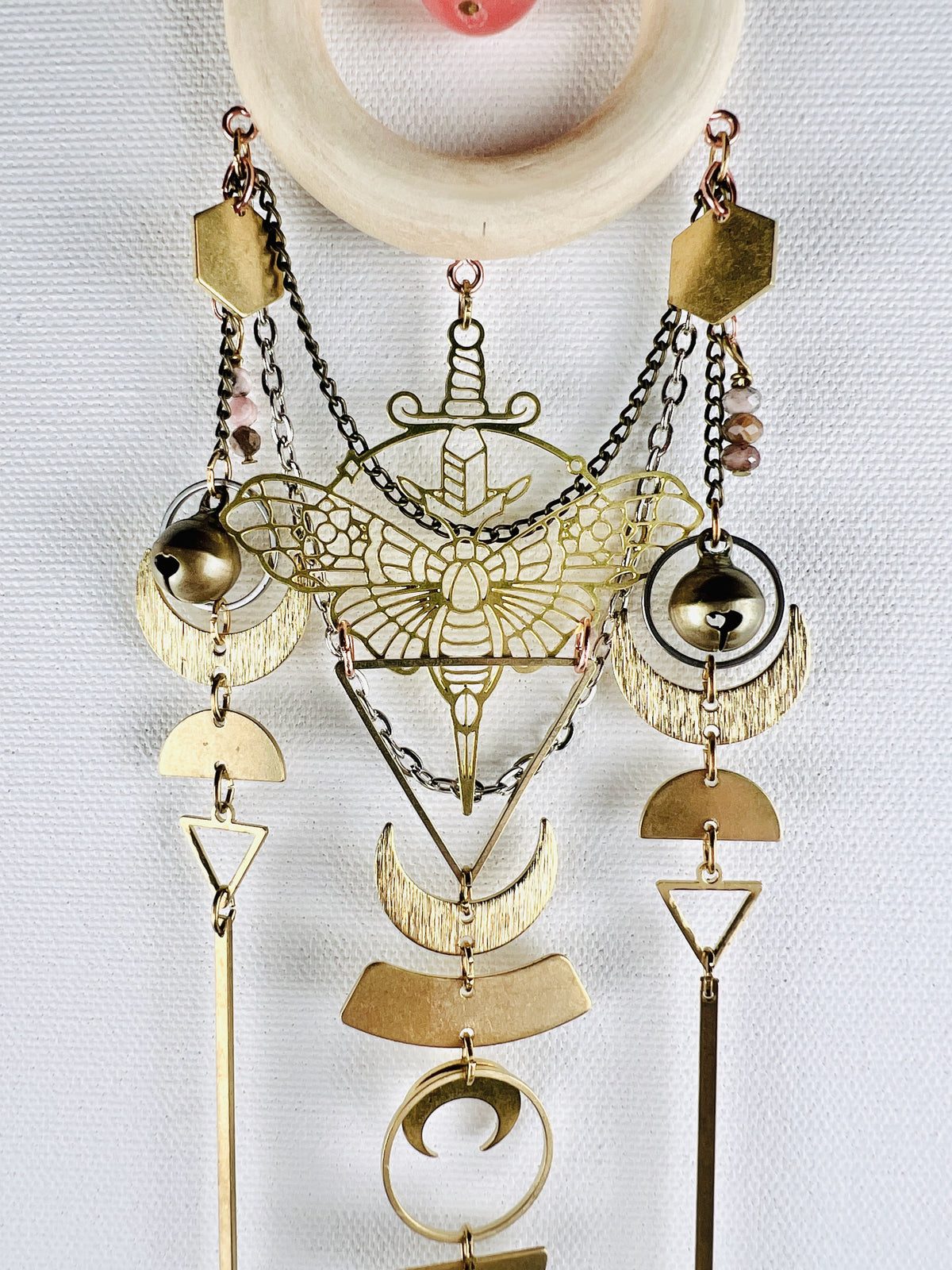 The bottom of a round pink bead, surrounded by a cream colored wood ring with attached components, such as: gold colored metal hexagons, a moth charm with sword, brass bells, silver and bronze chain, crescent moons, half circles, triangles, gold bars; all on a light gray campus wall.