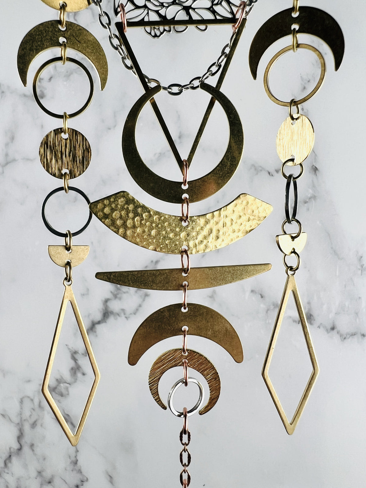 three sections of beautifully connected shapes of gold colored metal pieces, hanging vertically in front of a gray and white marble wall. Shapes on this piece include crescent moons, triangles, rings, circles, diamonds and copper and silver colored chain and are connected with small copper colored rings. 
