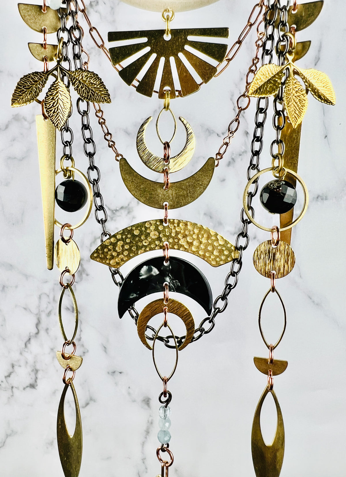 Depicted here is the bottom, middle section of a piece of hanging crystal wall art along with its black Tourmaline and aquamarine beads and metal components, which include half circles, leaves, triangles, circles, ovals, crescent moons - both metal and acrylic, and gun metal and copper colored chain. All hang beautifully patterned in front of a gray and white marble wall. The piece resembles a modern Dreamcatcher-like design.