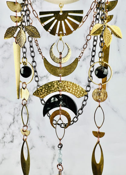 Depicted here is the bottom, middle section of a piece of hanging crystal wall art along with its black Tourmaline and aquamarine beads and metal components, which include half circles, leaves, triangles, circles, ovals, crescent moons - both metal and acrylic, and gun metal and copper colored chain. All hang beautifully patterned in front of a gray and white marble wall. The piece resembles a modern Dreamcatcher-like design.