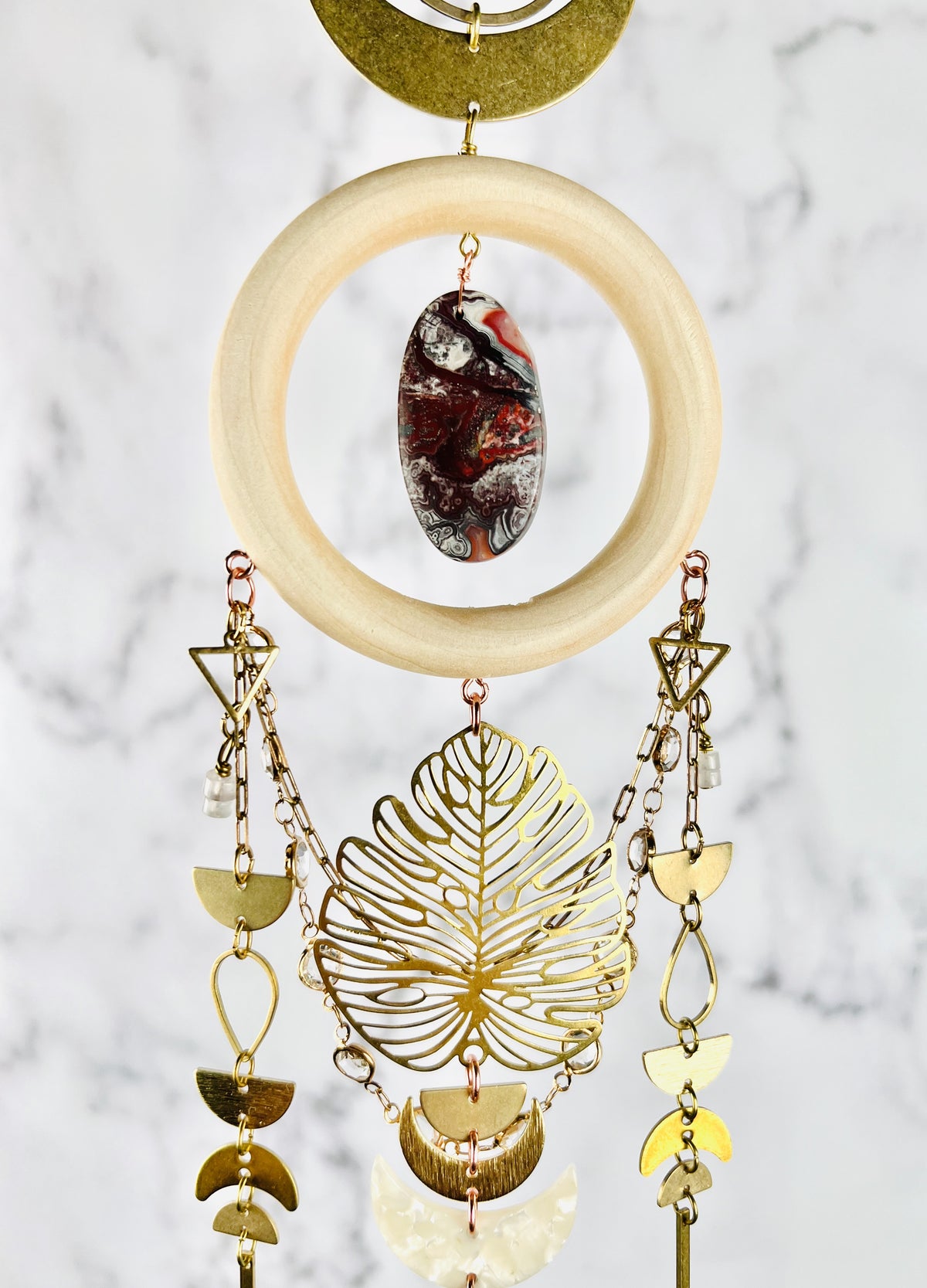 The bottom, half of dimensional hanging wall art, including: a bronze crescent, moon and ring, are connected to a cream colored wood ring with a center stone of red, black and gray, crazy lace agate. Connected to the bottom of the word ring are triangles, wire, wrapped beads, half circles, gold colored chain with prisms, teardrop shapes, crescent moons, and a large Monstera leaf. The peace hangs vertically in front of a gray and white marble wall.