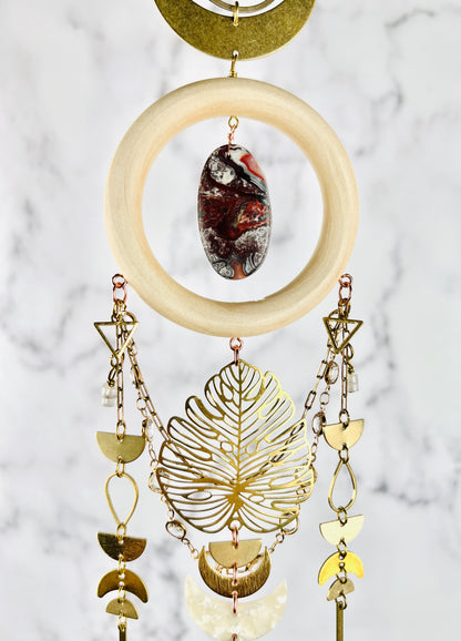 The bottom, half of dimensional hanging wall art, including: a bronze crescent, moon and ring, are connected to a cream colored wood ring with a center stone of red, black and gray, crazy lace agate. Connected to the bottom of the word ring are triangles, wire, wrapped beads, half circles, gold colored chain with prisms, teardrop shapes, crescent moons, and a large Monstera leaf. The peace hangs vertically in front of a gray and white marble wall.