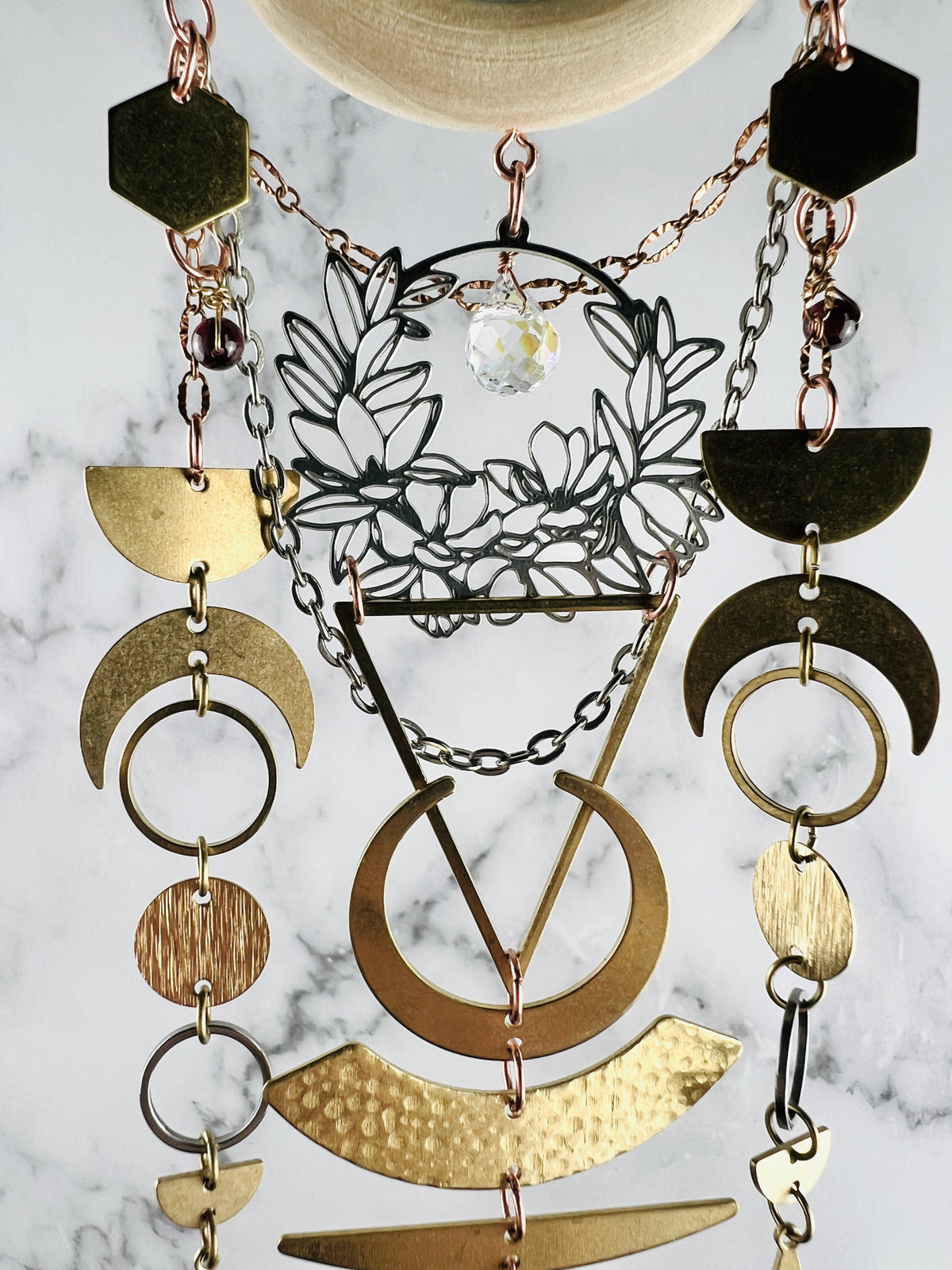 connected to a piece of light wood, hanging vertically are a silver colored metal wreath with a clear prism drop, silver and copper colored chain, eads hexagons, crescent moons, half moons, rings, circles and a large triangle. These components make a beautifully connected piece of wall art that hangs in front of a gray and white wall.