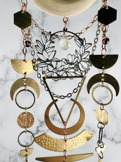 connected to a piece of light wood, hanging vertically are a silver colored metal wreath with a clear prism drop, silver and copper colored chain, eads hexagons, crescent moons, half moons, rings, circles and a large triangle. These components make a beautifully connected piece of wall art that hangs in front of a gray and white wall.