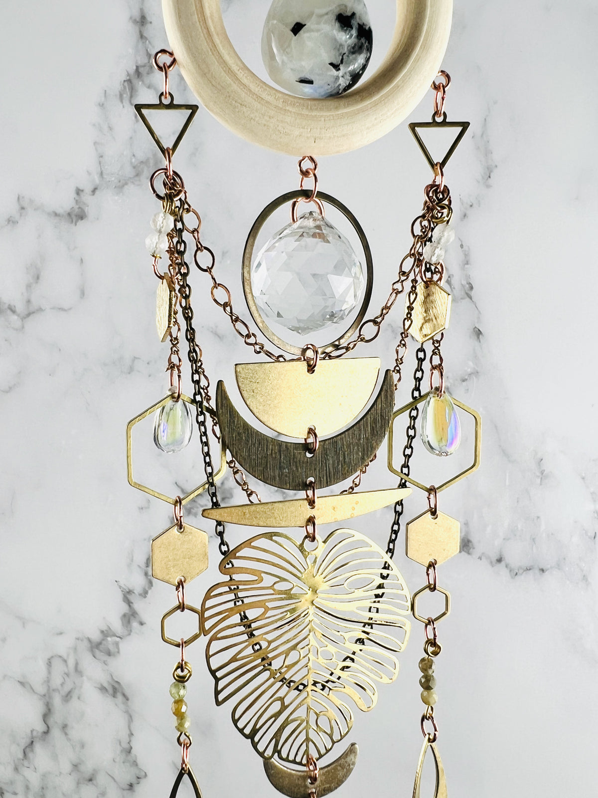 From the top of this crystal wall art, a wood ring surrounds a piece of moonstone. Below our various metal shapes and wire wrapped beads, including: triangles, prisms, rings, copper and bronze colored chain, hexagons, teardrops, crescent moons, half circle, green garnets and Monstera leaf; all hanging in front of a gray and white marble wall.