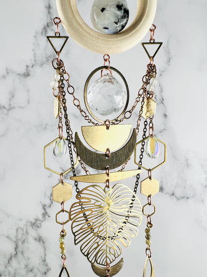 From the top of this crystal wall art, a wood ring surrounds a piece of moonstone. Below our various metal shapes and wire wrapped beads, including: triangles, prisms, rings, copper and bronze colored chain, hexagons, teardrops, crescent moons, half circle, green garnets and Monstera leaf; all hanging in front of a gray and white marble wall.
