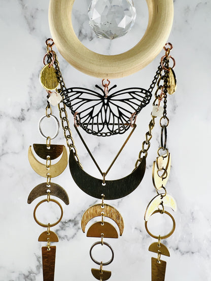 vertically hanging crystal wall art with metal charms including a center silver butterfly, crescent moons, half circles, triangle, silver and gold rings, chain and crystals on a marble background. Pink Aventurine with large prism in the center of a light wood ring. A suncatcher and manifestation charm, created for intentional home decor and healing.