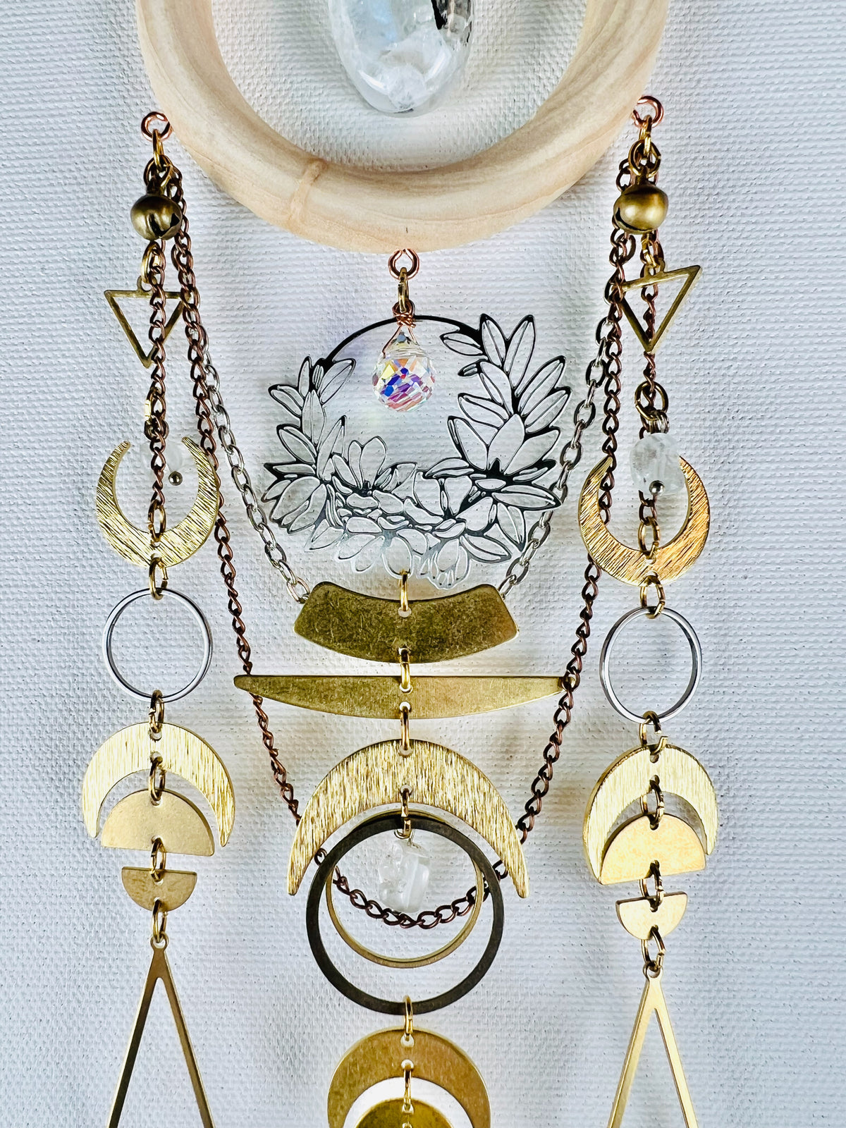A light wood ring surrounding an oval moonstone hangs at the top, connected to this are bronze and silver chains, Rings and gold colored metal triangles, bells, crescent moons, half circles and wire wrapped beads. This crystal art hangs on a light gray canvas wall.