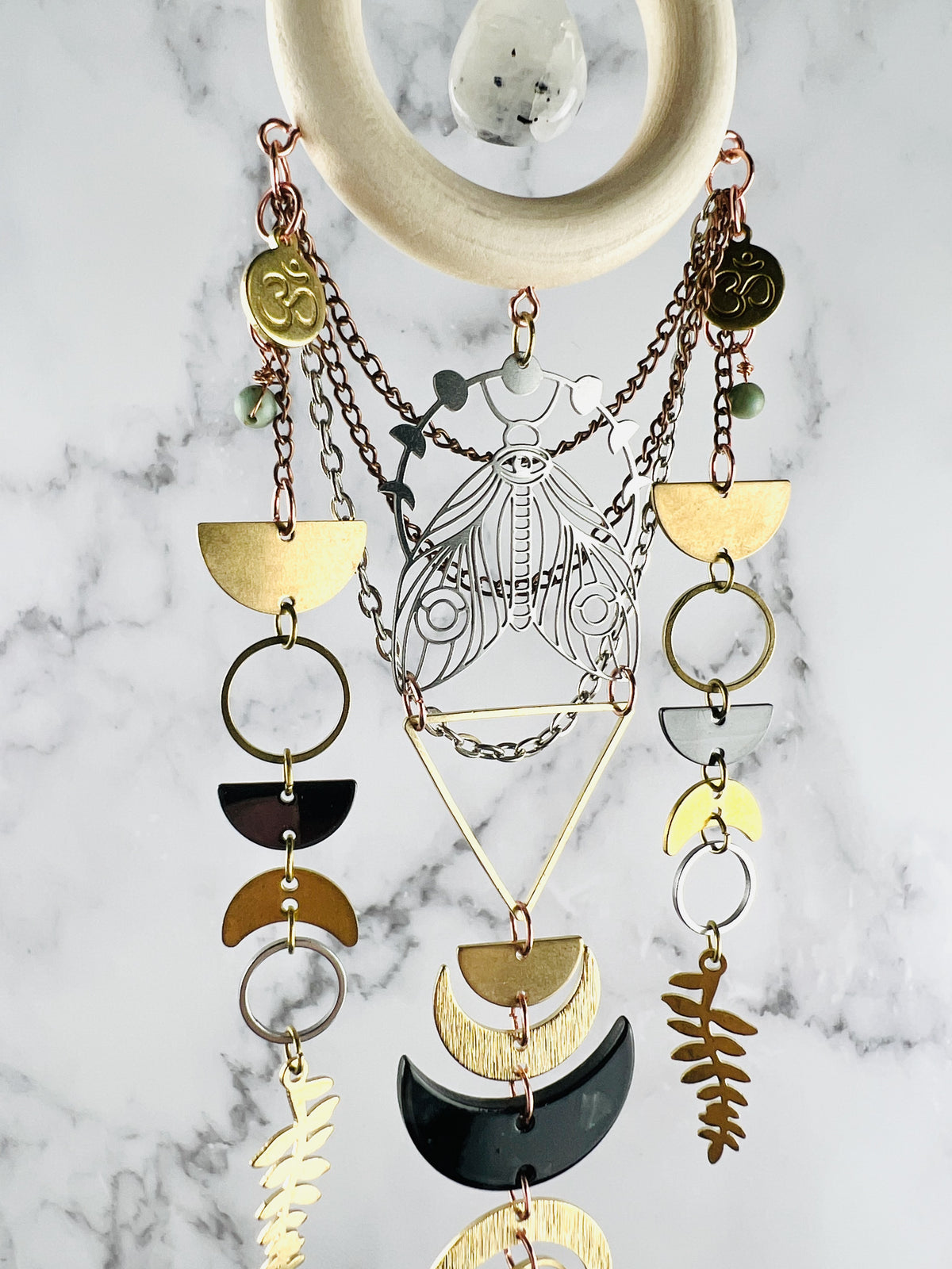 A vertically hanging piece of crystal wall art, with components connected by tiny rings, including: a moonstone, a cream colored wood ring, Ohm symbols, wire wrapped light, blue beads, silver and bronze colored chain, half circles, silver and gold rings, crescent moons, fern leaves and a silver moth charm with moon faces above it. The entire piece hangs in front of a gray and white marble wall.