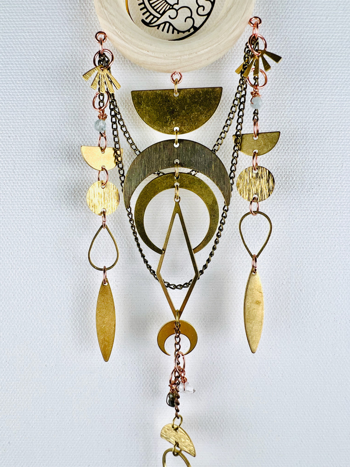 A hanging piece of crystal wall art, including wire wrapped light, blue beads, a light colored wood, ring, bronze chains and gold colored metal shapes connected by tiny copper metal rings. Elements on this piece include fans, half circles, Crescent moons, diamonds, ovals, teardrops. Chains swoop in the background of three separate combinations of the above mentioned items formed into beautiful vertical patterns. It hangs on a light grey background.
