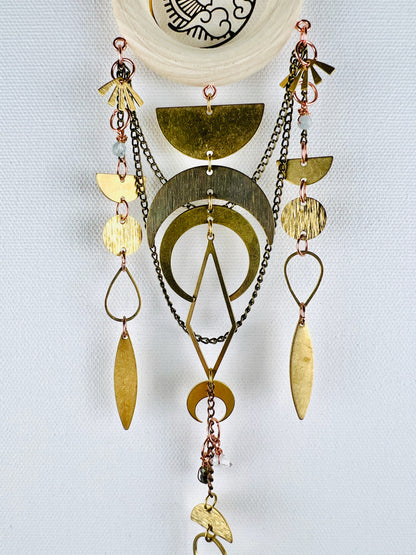 A hanging piece of crystal wall art, including wire wrapped light, blue beads, a light colored wood, ring, bronze chains and gold colored metal shapes connected by tiny copper metal rings. Elements on this piece include fans, half circles, Crescent moons, diamonds, ovals, teardrops. Chains swoop in the background of three separate combinations of the above mentioned items formed into beautiful vertical patterns. It hangs on a light grey background.