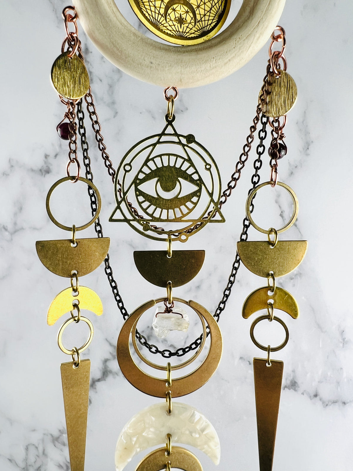 middle view of Evil eye symbol surrounded by triangle and rings with Wood ring, celestial Charm, gold medal Components, Including Rings, evil eye, crescent, moons, half circles, long triangles with wire wrapped beads and acrylic crescent moon with chain connected to it; all hanging on a marble wall.
