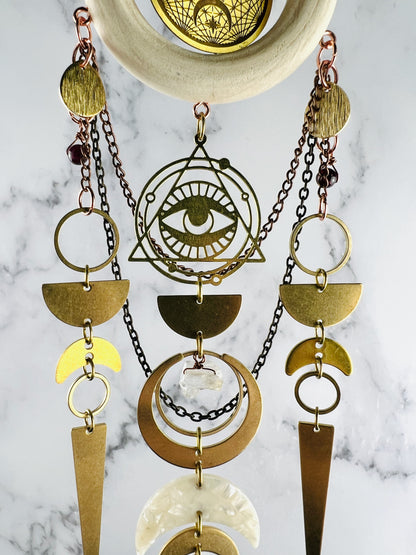middle view of Evil eye symbol surrounded by triangle and rings with Wood ring, celestial Charm, gold medal Components, Including Rings, evil eye, crescent, moons, half circles, long triangles with wire wrapped beads and acrylic crescent moon with chain connected to it; all hanging on a marble wall.