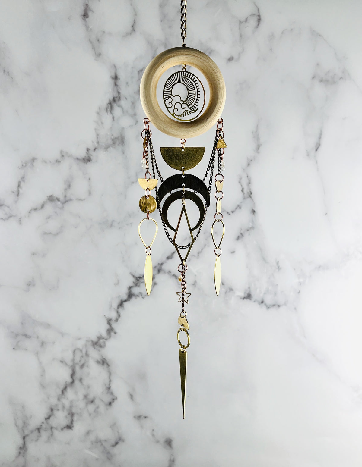 Decorative vertically hanging wall art with crystals and geometric metal designs on a marble background. Sun & cloud charm with half circles, circles, crescent moons, diamonds, and triangles with wood ring hanging in front of a marble wall. Handcrafted Moonstone and Citrine manifestation charm wall art created for intentional home decor and emotional balance.