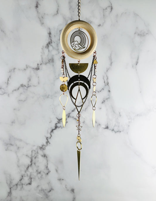 Decorative vertically hanging wall art with crystals and geometric metal designs on a marble background. Sun & cloud charm with half circles, circles, crescent moons, diamonds, and triangles with wood ring hanging in front of a marble wall. Handcrafted Moonstone and Citrine manifestation charm wall art created for intentional home decor and emotional balance.