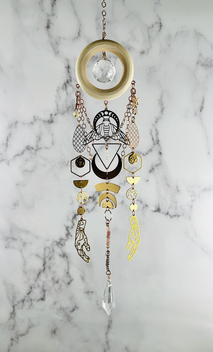 Decorative hanging crystal wall art with geometric designs on a marble background. wood, golden and silver metal charms including hands, moon phase and a bee with hexagons, half circles and om symbols. prism at the bottom
