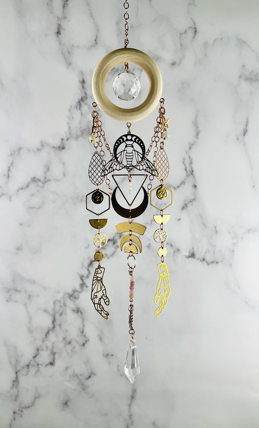 Decorative hanging crystal wall art with geometric designs on a marble background. wood, golden and silver metal charms including hands, moon phase and a bee with hexagons, half circles and om symbols. prism at the bottom