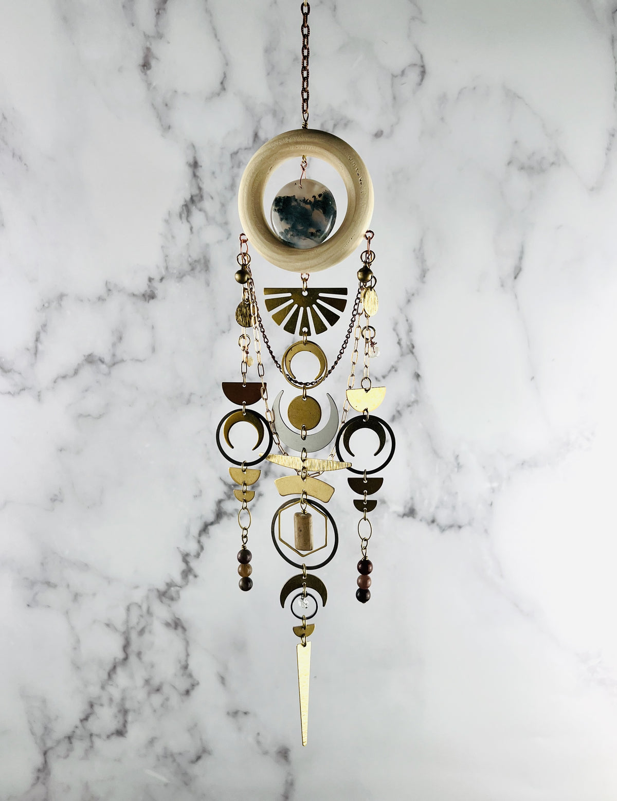 Hanging crystal wall art made of a wood ring with center stone of moss agate. Dangling from the ring are gold and silver colored metal shapes including: a sunburst, bells, crescent moons, half circles, rings ovals, hexagons, triangles and wire wrapped Topaz, Petrified Wood, Jasper & Herkimer Diamond beads. suspended from a gold colored chain in front of a marble background.
