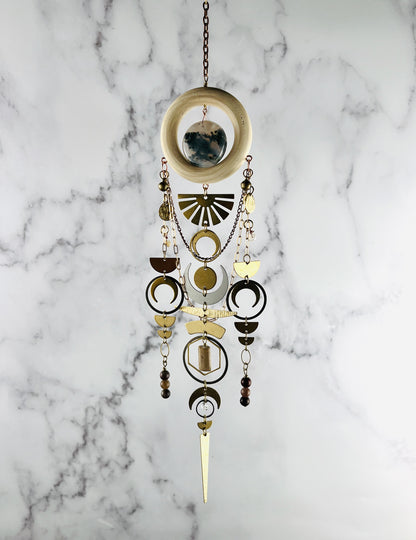 Hanging crystal wall art made of a wood ring with center stone of moss agate. Dangling from the ring are gold and silver colored metal shapes including: a sunburst, bells, crescent moons, half circles, rings ovals, hexagons, triangles and wire wrapped Topaz, Petrified Wood, Jasper & Herkimer Diamond beads. suspended from a gold colored chain in front of a marble background.