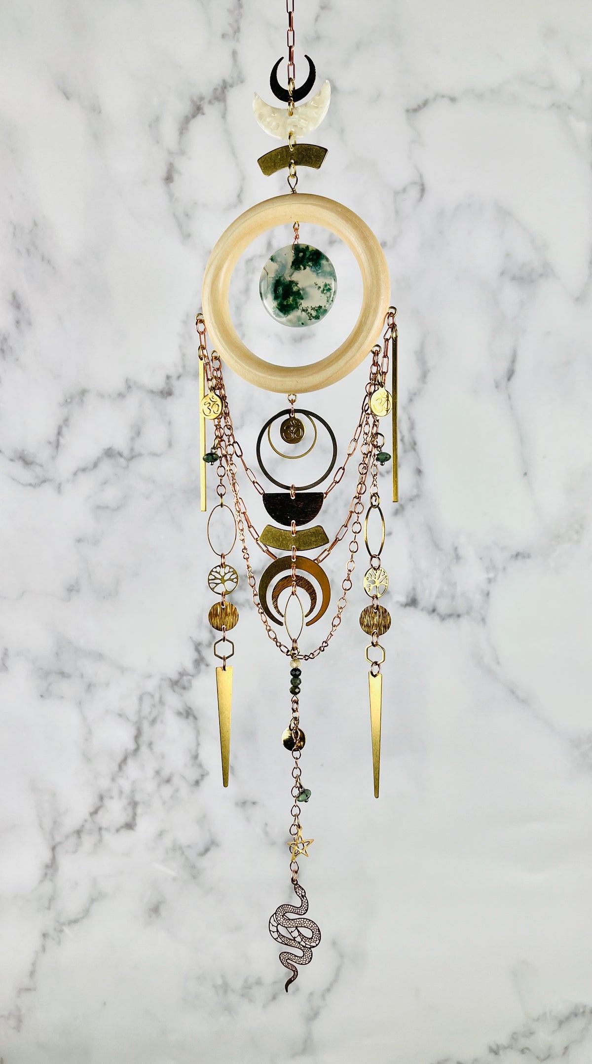 A piece of crystal wall art hangs in front of a marble wall. The Charm features copper colored chain, crescent moons, a light wood ring with a piece of green and translucent disk of moss agate, at its center. Below hang more attached copper colored chains with connected gold colored metal moons, tree of life, ohm symbol, wire wrapped beads, triangles, hexagons, ovals, stars, and a metal snake charm at the bottom.