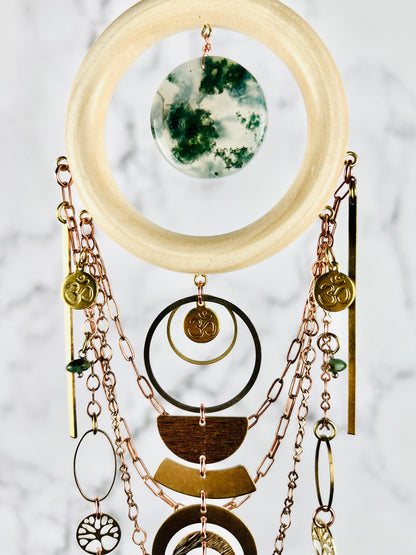 Middle portion of a crystal hanging wall art piece made of wood ring, moss agate, Home symbols, metal components, including copper colored chain, long dangling bars, ovals, rings, half circles and tree of life, hanging in front of a marble wall.