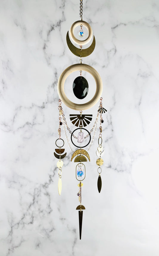 Decorative hanging ornament with wood, geometric shapes and crystals on a marble background. Handmade Obsidian, Bloodstone, and Amethyst wall art manifestation charm with chains, circles, moons, half-circles, triangles and elongated triangles, created for intentional home decor and emotional grounding.
