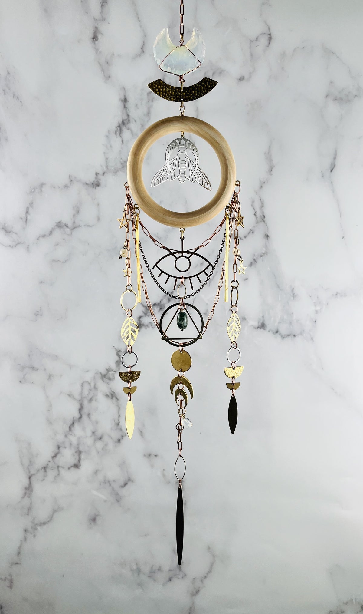Decorative wall hanging crystal wall art with geometric patterns and beads on a marble background. vertically hanging featuring gold colored metal crescent moons, moon phase, silver bee, stars, triangle, rings, evil eye protection symbol, golden leaves, various stones, and circles.