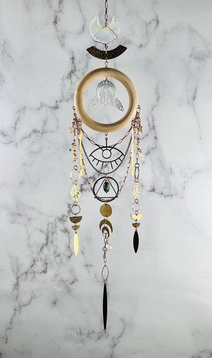 Decorative wall hanging crystal wall art with geometric patterns and beads on a marble background. vertically hanging featuring gold colored metal crescent moons, moon phase, silver bee, stars, triangle, rings, evil eye protection symbol, golden leaves, various stones, and circles.