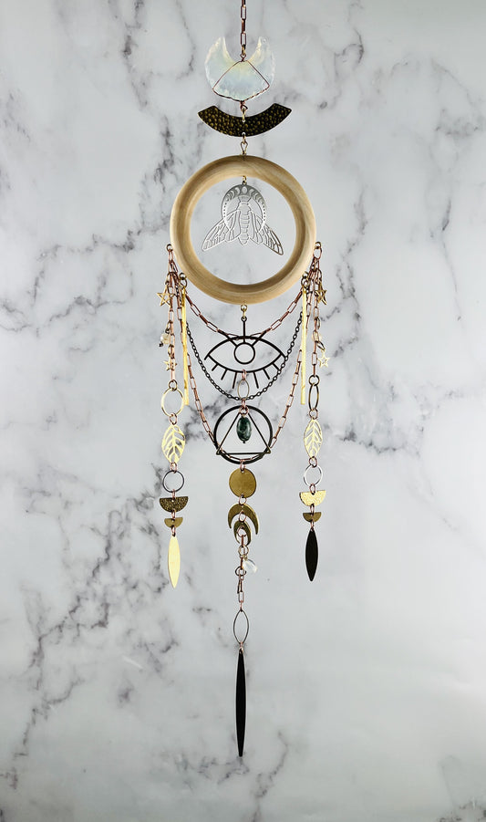 Decorative wall hanging crystal wall art with geometric patterns and beads on a marble background. vertically hanging featuring gold colored metal crescent moons, moon phase, silver bee, stars, triangle, rings, evil eye protection symbol, golden leaves, various stones, and circles.