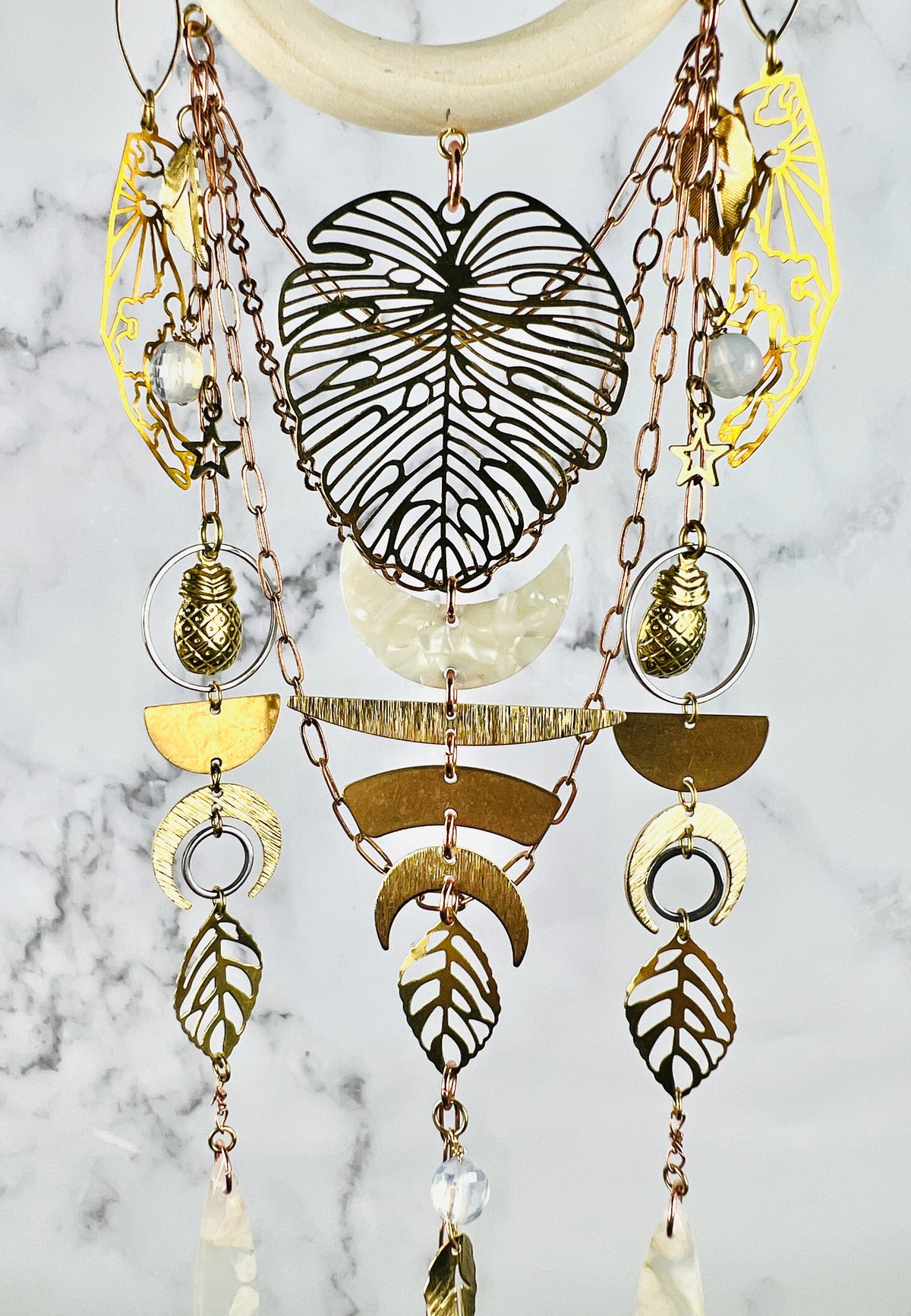 Dangling from a wood ring, hangs various metal components, including hands, a large Monstera leaf, Opalite crystals, pineapple, charms, copper colored chain, a cream colored crescent moon, as well as metal, crescent, moon shapes, stainless steel rings, leaves, and flower agate beads. The piece resembles a Dreamcatcher and hangs beautifully in front of a gray and white marble wall.