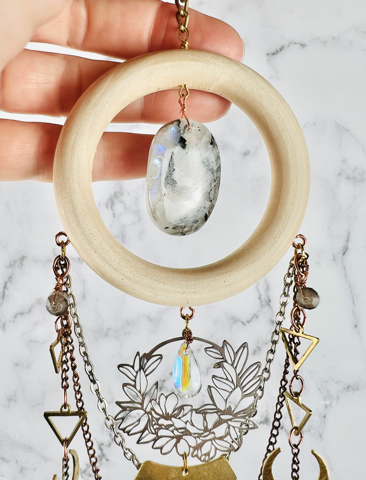 A hand holding a bohemian-style dreamcatcher like crystal wall art piece featuring a light natural wood ring, a polished moonstone pendant, and dangling chains with geometric and floral metal charms against a gray and white marble background.