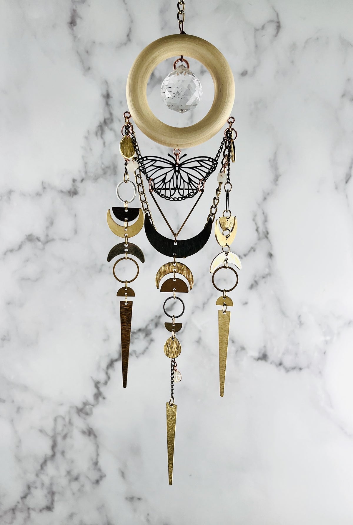 vertically hanging crystal wall art with metal charms including a silver butterfly, crescent moons, half circles, triangles, rings, chain and crystals on a marble background. Pink Aventurine and Topaz with large prism in the center of a light wood ring. A suncatcher and manifestation charm, created for intentional home decor and emotional balance.