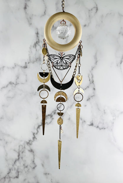 vertically hanging crystal wall art with metal charms including a silver butterfly, crescent moons, half circles, triangles, rings, chain and crystals on a marble background. Pink Aventurine and Topaz with large prism in the center of a light wood ring. A suncatcher and manifestation charm, created for intentional home decor and emotional balance.