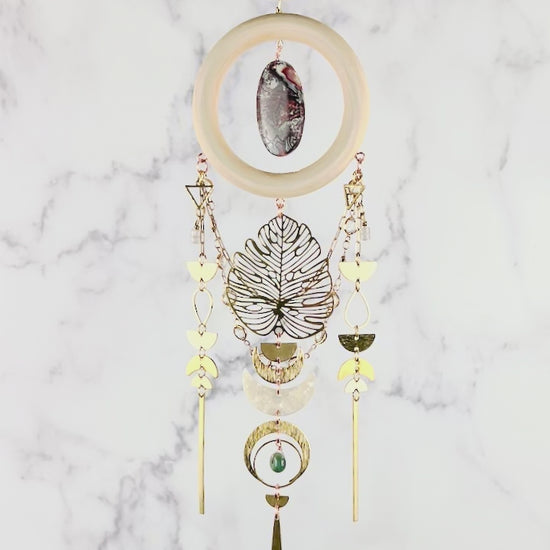 Gently spinning, vertically hanging hypersthene, red and gray crazy lace agate, rose quartz, and Moss agate crystal wall art. Gold chain suspense, a dimensional piece of art, including connected elements of metal, cream colored wood rings, and crystals. Featuring, gold colored metal rings, crescent moons, triangles, half circles, tear Drop shapes, a Monstera leaf and long gold bars, connected with tiny rings. The piece hangs in front of a gray and white marble wall.