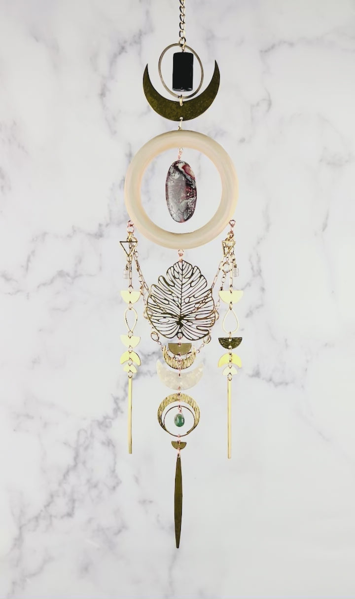 Gently spinning, vertically hanging hypersthene, red and gray crazy lace agate, rose quartz, and Moss agate crystal wall art. Gold chain suspense, a dimensional piece of art, including connected elements of metal, cream colored wood rings, and crystals. Featuring, gold colored metal rings, crescent moons, triangles, half circles, tear Drop shapes, a Monstera leaf and long gold bars, connected with tiny rings. The piece hangs in front of a gray and white marble wall.