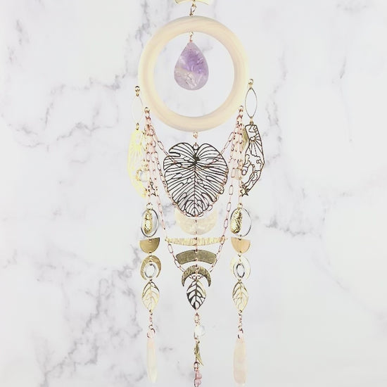 peacefully spinning vertical crystal wall art consisting of stones including: obsidian, Opalite, flower agate, and lepidolite plus copper colored chain, a light wood ring, a teardrop shaped purple amethyst, and various gold metal charms including: a large monstera leaf, pineapples, Half circles, crescent, moons, leaves, and beads, hang in front of a marble wall.