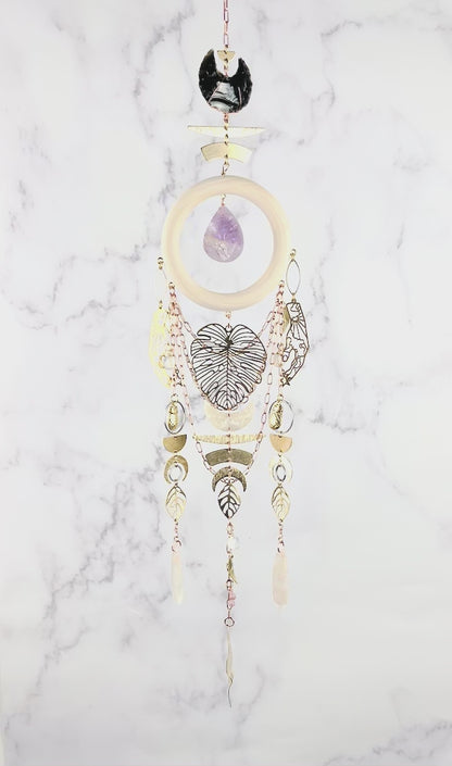 peacefully spinning vertical crystal wall art consisting of stones including: obsidian, Opalite, flower agate, and lepidolite plus copper colored chain, a light wood ring, a teardrop shaped purple amethyst, and various gold metal charms including: a large monstera leaf, pineapples, Half circles, crescent, moons, leaves, and beads, hang in front of a marble wall.