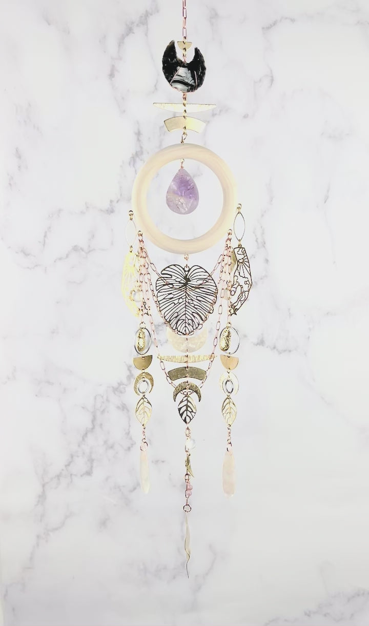peacefully spinning vertical crystal wall art consisting of stones including: obsidian, Opalite, flower agate, and lepidolite plus copper colored chain, a light wood ring, a teardrop shaped purple amethyst, and various gold metal charms including: a large monstera leaf, pineapples, Half circles, crescent, moons, leaves, and beads, hang in front of a marble wall.