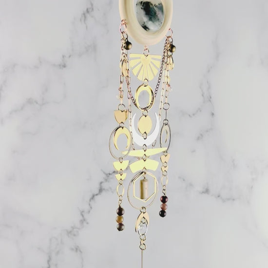 Vertically hanging, crystal wall art piece, consisting of moss agate, topaz, petrified wood, Jasper, Herkimer, diamond, moons, rings, circles, half circles, a sunburst, chain and light colored wood ring. All spin gently in front of a marble wall.