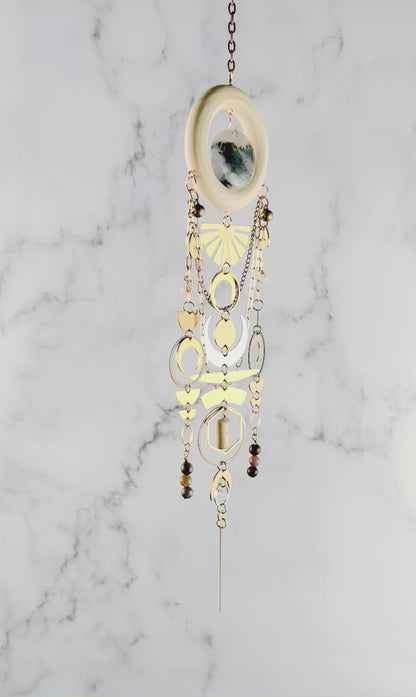 Vertically hanging, crystal wall art piece, consisting of moss agate, topaz, petrified wood, Jasper, Herkimer, diamond, moons, rings, circles, half circles, a sunburst, chain and light colored wood ring. All spin gently in front of a marble wall.