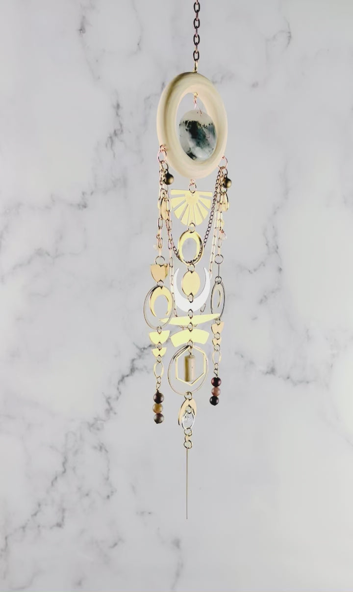 Vertically hanging, crystal wall art piece, consisting of moss agate, topaz, petrified wood, Jasper, Herkimer, diamond, moons, rings, circles, half circles, a sunburst, chain and light colored wood ring. All spin gently in front of a marble wall.