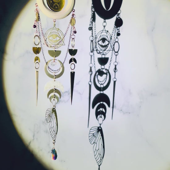 spotlight shining on a celestial wall art with crystals, light, wood ring, metal gold colored eye charm with triangle and Rings around it, bronze, colored chain, wire, wrapped beads, rings, half circles, crescent moons, citrine, prism, and a bug wing slowly sparkling and spinning, hanging from a chain in front of a marble wall.
