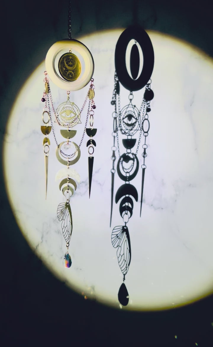 spotlight shining on a celestial wall art with crystals, light, wood ring, metal gold colored eye charm with triangle and Rings around it, bronze, colored chain, wire, wrapped beads, rings, half circles, crescent moons, citrine, prism, and a bug wing slowly sparkling and spinning, hanging from a chain in front of a marble wall.