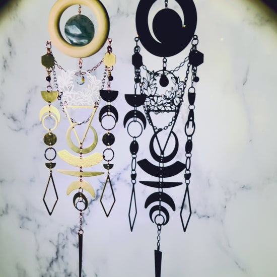 a piece of vertically hanging, crystal wall art with moss agate and garnet spins slowly, sparkling, surrounded by a spotlight, in front of a gray and white marble wall. This piece includes: bronze colored chains, a light wood ring, a round moss agate stone. Connected to the bottom of the wood, hanging below are gold and silver colored charms including: hexagons, half and crescent moons, rings, circles, diamonds and triangles; all connected by tiny rings.