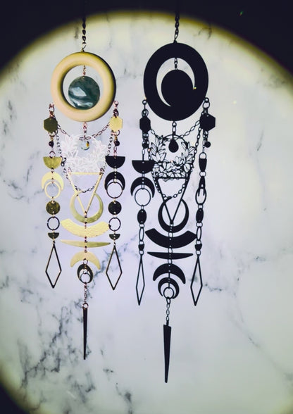 a piece of vertically hanging, crystal wall art with moss agate and garnet spins slowly, sparkling, surrounded by a spotlight, in front of a gray and white marble wall. This piece includes: bronze colored chains, a light wood ring, a round moss agate stone. Connected to the bottom of the wood, hanging below are gold and silver colored charms including: hexagons, half and crescent moons, rings, circles, diamonds and triangles; all connected by tiny rings.