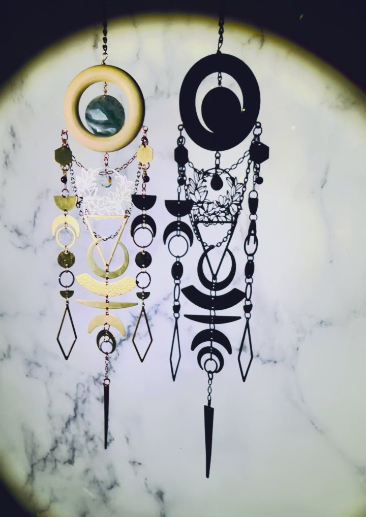a piece of vertically hanging, crystal wall art with moss agate and garnet spins slowly, sparkling, surrounded by a spotlight, in front of a gray and white marble wall. This piece includes: bronze colored chains, a light wood ring, a round moss agate stone. Connected to the bottom of the wood, hanging below are gold and silver colored charms including: hexagons, half and crescent moons, rings, circles, diamonds and triangles; all connected by tiny rings.