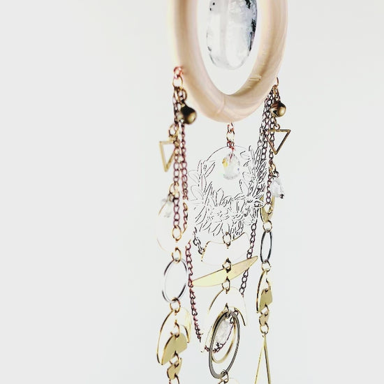 A piece of crystal wall art slowly spins in front of a white surface. This piece includes vertically hanging patterns of gold colored and silver colored metal charms such as: triangles, bells, chain, a flower wreath, circles, crescent moons, half moons, diamond shapes, and a snake Charm.